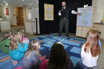 young numismatists sitting in a circle on the floor, looking up at a speaker